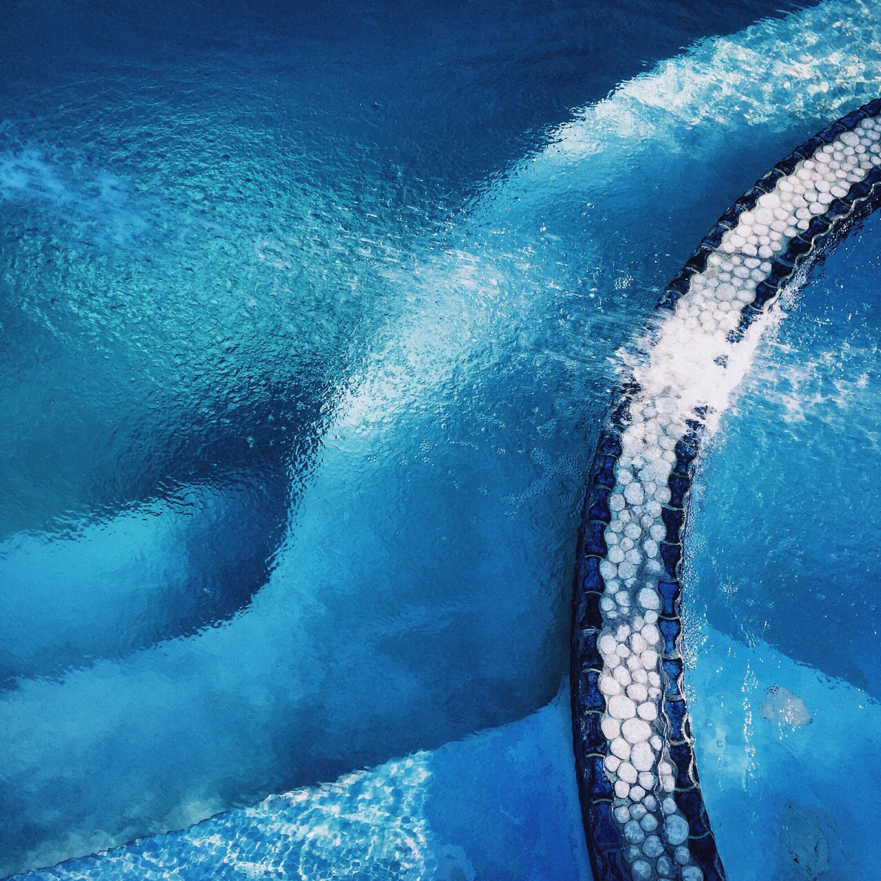 Overhead view of rippling blue pool water with textured tiled edge, cool and serene