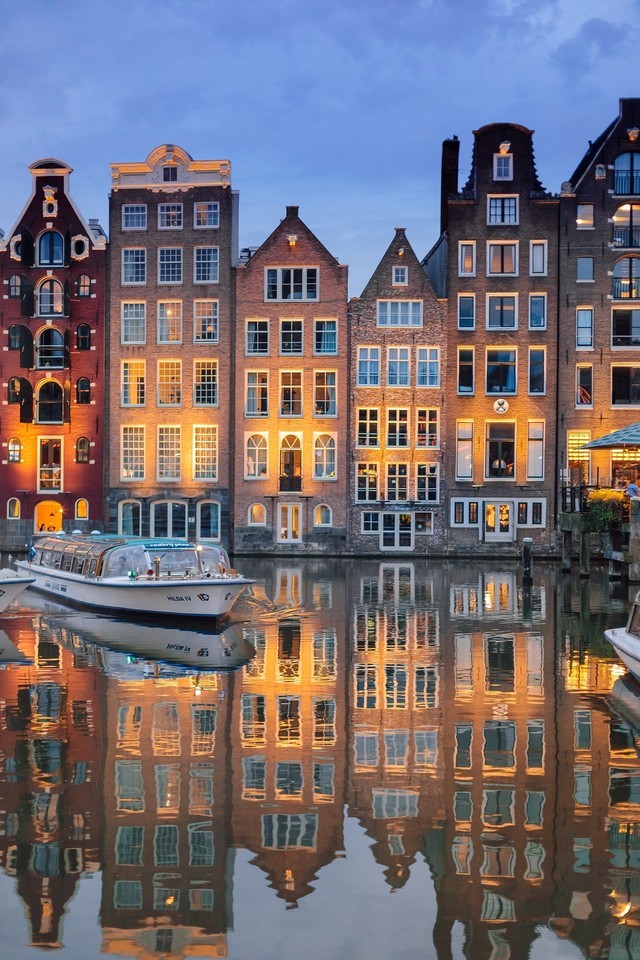Row of tall narrow European canal houses illuminated at night, reflected in calm water, warm amber lighting
