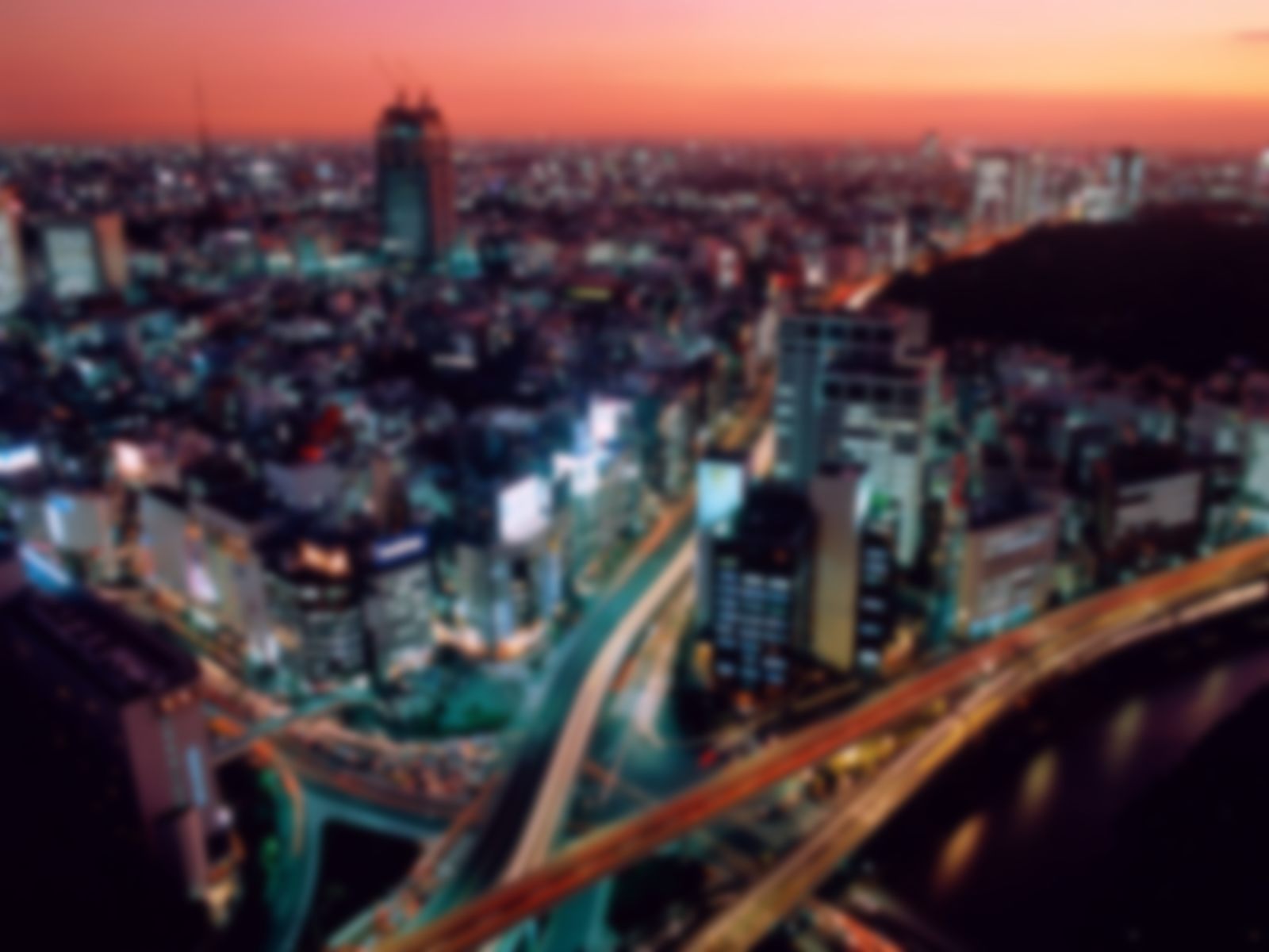 Blurred aerial night cityscape with illuminated buildings and elevated highways, pink-orange sunset sky